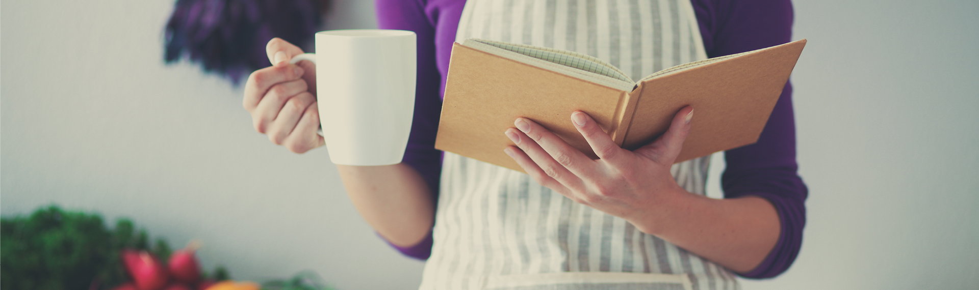 Eine weibliche Person steht mit einer Tasse in der rechten Hand und einem Kochbuch in ihrer linken Hand in der Küche.